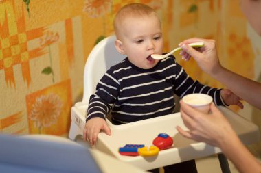 Aile hayatı vurulması. Seçici odaklanma. Sıcak dergi tonu, fazladan ses. Annem bebeği bir kaşık yoğurtla sandalyede besliyor.