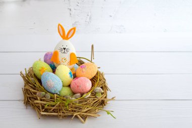 Beautiful multi-colored decorated Easter eggs and a cute Easter bunny in a nest of hay and pussy willow on a white wooden background. Ideas for decorating for the holiday. Copy space.Happy easter