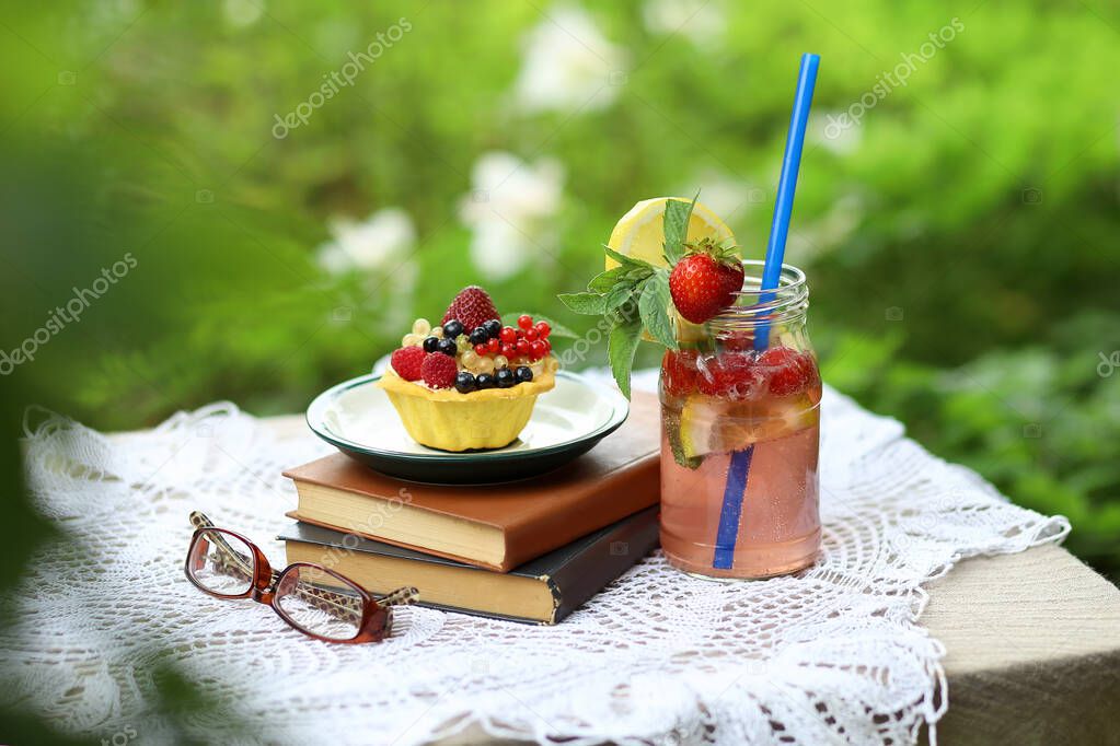 Homemade strawberry mojito lemonade in a glass jar with a blue straw, reading glasses and a cupcake with summer berries on a stack of books on a table in the garden. Summer leisure