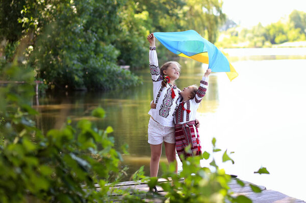 Girls sister friends in embroidered national Ukrainian costumes with a yellow-blue flag on a pier on the shore of the lake. Independence day of ukraine, constitution, vyshyvanka day, flag day