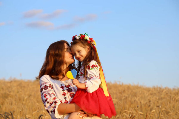 Happy smiling Ukrainian Mom and daughter in vyshyvanka (embroidered shirts) with a yellow-blue heart on a background of wheat field and sky.Ukraine's Independence Day. National Flag Day of Ukraine