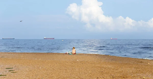 温かみのある晴れた夏の日 彼女の肩とジーンズのショート パンツに吟遊詩人の少女の悲しい海岸にカメラに背を向けて座っているし 海を見て — ストック写真