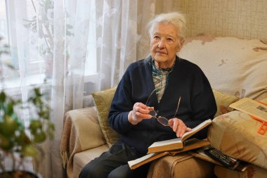 Evde rahat bir koltukta kitap okuyan yaşlı bir kadın.