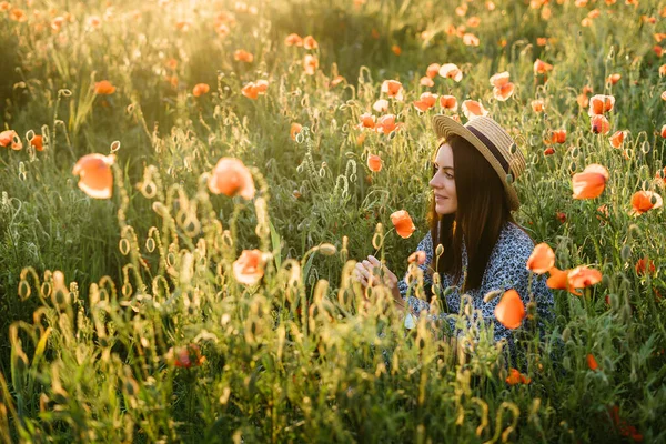 ポピーの分野でポーズをとっている帽子と青いドレスの素晴らしい若い女性 夕暮れ時の夏の野花の香りを楽しむ美しい優しい若い女性 — ストック写真