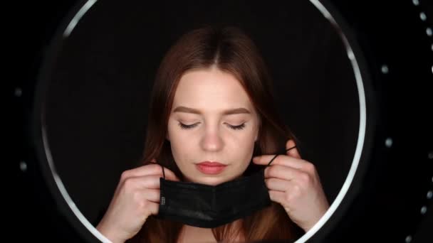 A beautiful girl on a black background wears a black mask illuminated by a beautiful circular light looks into the camera. Master in a beauty salon preparing for the procedure — Stock Video