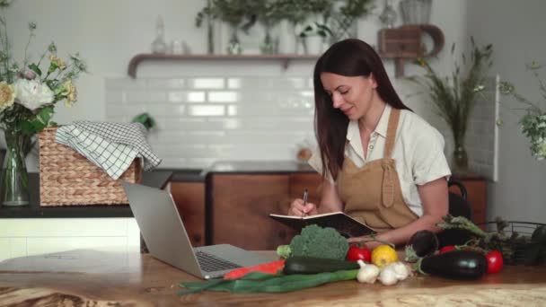 一位年轻漂亮的女士从新鲜的有机蔬菜中准备了一道菜谱，她看着厨房里的笔记本电脑屏幕，在笔记本里写下了一道菜谱，她穿着亚麻布衣服。学习网上营养学 — 图库视频影像