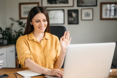 Genç, güzel bir kadın video odasında konuşuyor ve ana ofiste masada otururken el sallıyor.