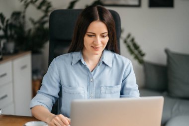 Evde bilgisayarla çalışan mutlu ve serbest bir genç kadın. Çevrimiçi öğrenim gören çekici iş kadını, dizüstü bilgisayar, web sörfü bilgileri veya internet mağazasından alışveriş yapıyor..