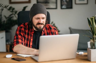 Odaklanmış sakallı genç adam, günlük giysiler içinde dizüstü bilgisayar kullanıyor, klavyeye yazıyor, e-posta ya da mesaj yazıyor, sohbet ediyor, alışveriş yapıyor, bilgisayar üzerinde çalışan başarılı bir serbest yazar. 