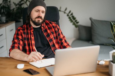 Odaklanmış sakallı genç adam, günlük giysiler içinde dizüstü bilgisayar kullanıyor, klavyeye yazıyor, e-posta ya da mesaj yazıyor, sohbet ediyor, alışveriş yapıyor, bilgisayar üzerinde çalışan başarılı bir serbest yazar. 