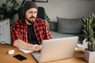 Odaklanmış sakallı genç adam, günlük giysiler içinde dizüstü bilgisayar kullanıyor, klavyeye yazıyor, e-posta ya da mesaj yazıyor, sohbet ediyor, alışveriş yapıyor, bilgisayar üzerinde çalışan başarılı bir serbest yazar. 