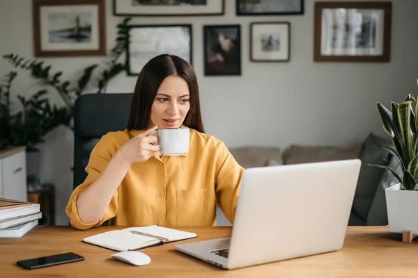 Hermosa mujer morena joven en ropa amarilla casual usando el ordenador portátil mientras trabaja en casa bebe café — Foto de Stock