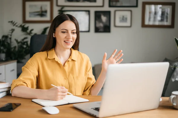 Joven bonita mujer hablando en videollamada y saludando la mano mientras está sentada en la mesa en la habitación de la oficina — Foto de Stock