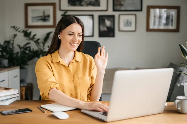 Joven bonita mujer hablando en videollamada y saludando la mano mientras está sentada en la mesa en la habitación de la oficina — Foto de Stock