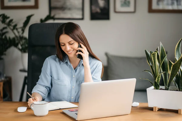 Mujer de negocios sonriente hablando en un teléfono móvil y utilizando un ordenador portátil mientras trabaja en casa, mirando a la pantalla, leyendo un mensaje, una joven empleada aconseja a un cliente, conversaciones telefónicas — Foto de Stock