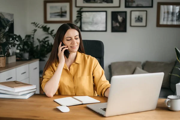 Mujer de negocios sonriente hablando en un teléfono móvil y utilizando un ordenador portátil mientras trabaja en casa, mirando a la pantalla, leyendo un mensaje, una joven empleada aconseja a un cliente, conversaciones telefónicas — Foto de Stock