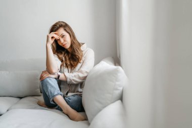 Genç bayan oturma odasındaki kanepede oturuyor. Beyaz rahat ev kıyafetleri içinde güzel bir sarışın.