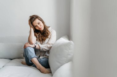 Genç bayan oturma odasındaki kanepede oturuyor. Beyaz rahat ev kıyafetleri içinde güzel bir sarışın.
