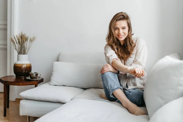 Uma jovem loira em roupas casuais e confortáveis senta-se em um sofá branco em casa — Fotografia de Stock