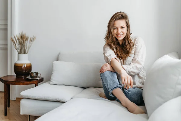 A jovem está sentada no sofá da sala de estar. Uma bela loira em branco roupas confortáveis para casa — Fotografia de Stock