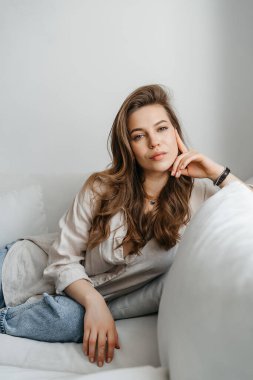 Rahat giysiler içinde genç sarışın bir kadın evdeki beyaz koltukta oturuyor.