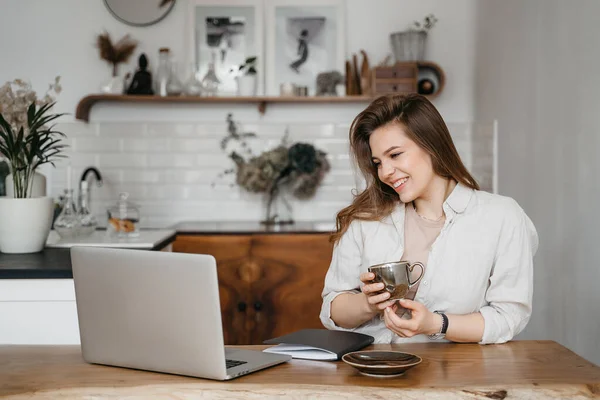 Jovem atraente feliz sentada em sua cozinha no balcão com um laptop sorrindo para a câmera — Fotografia de Stock