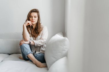 Genç bayan oturma odasındaki kanepede oturuyor. Beyaz rahat ev kıyafetleri içinde güzel bir sarışın.