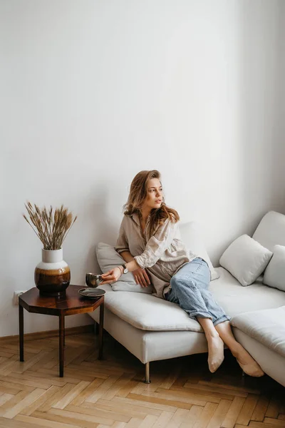 Uma jovem loira em roupas casuais e confortáveis senta-se em um sofá branco em casa e bebe café. interior escandinavo, minimalismo — Fotografia de Stock