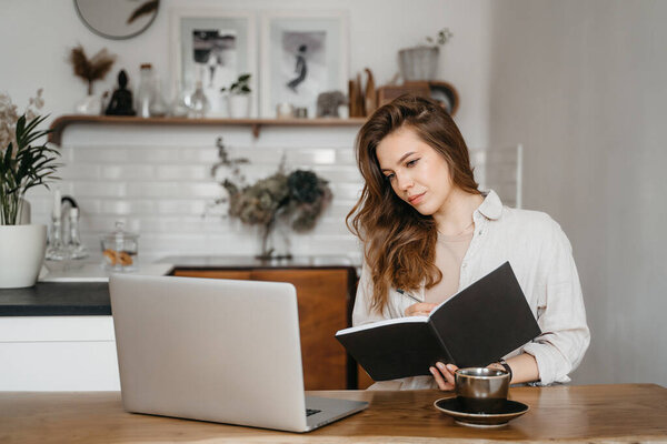 Smiling young pretty woman office worker business lady listening to an online webinar on laptop, taking notes in notebook, working on freelance project at home, remote work, learning at home concept