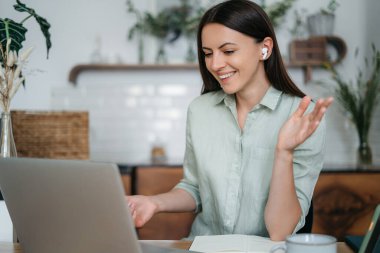 Genç, güzel bir kadın telefonda konuşuyor ve mutfakta masada otururken el sallıyor.