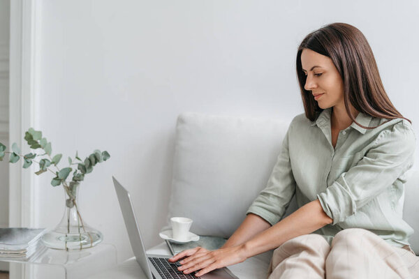 Сосредоточенная молодая женщина сидит на диване в гостиной и работает вдали от ноутбука. Тысячелетний женский отдых на диване на домашнем обучении онлайн, просмотр или серфинг беспроводной интернет на гаджете.