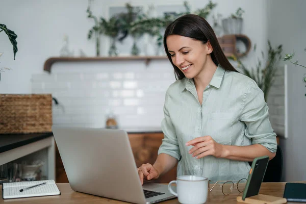 Jong mooi vrouw praten op video oproep en zwaaien hand terwijl zitten aan tafel in de keuken — Stockfoto