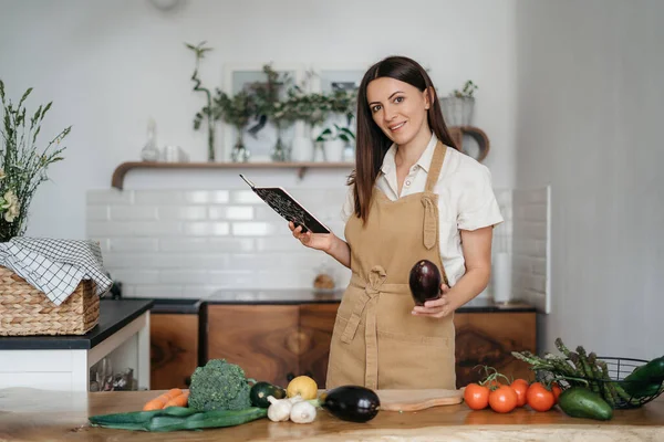 Bela jovem morena mulher se prepara na cozinha de acordo com uma receita de um notebook uma refeição saudável de vegetais orgânicos frescos olha para a câmera — Fotografia de Stock