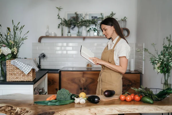 Bela jovem morena prepara uma refeição saudável de vegetais orgânicos frescos na cozinha de acordo com uma receita de um caderno — Fotografia de Stock