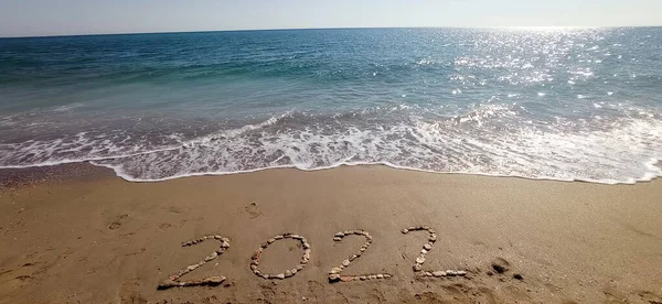 Mutlu yıllar 2022, plajda el sallayarak ve açık mavi denizle. Deniz kıyısında 2022 yılına ait sayılar. Güzel plaj arka planında altın kumda el yazısıyla yazılmış mesaj. Yeni yıl kavramı.