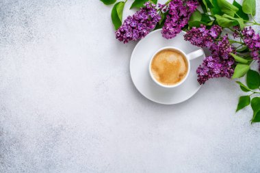 Sütlü bir fincan taze kahve ve desenli gri arka planda yeşil yapraklı çiçek açan mor leylak dalı. Kahve molası. Bahar çiçekleri ve köpüklü kapuçino..
