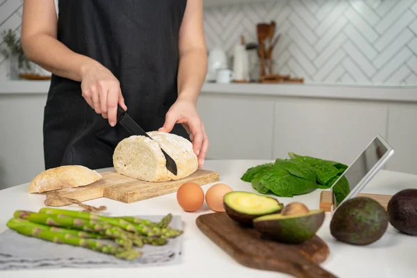 Instruction, step 1. Ingredients for making toasts for healthy breakfast. Woman cutting bread. Avocado on wooden board, asparagus on napkin, eggs, romano salad and tablet with recipe.