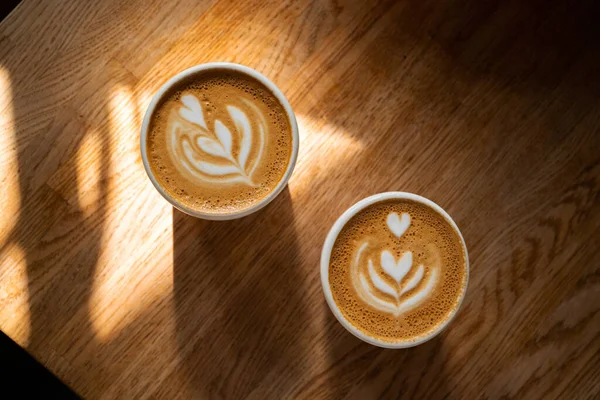 Dua cangkir kopi segar dengan seni latte di atas meja kayu di bawah sinar matahari. Pagi cappuccino dengan hati di kafe untuk pasangan. Konsep coffee break. Minuman kafein untuk sarapan tutup. Tampilan atas, berbaring datar — Stok Foto