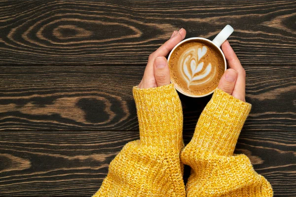 Wanita tangan di sweter kuning memegang cangkir kopi segar dengan seni latte yang indah di latar belakang kayu. Selamat musim gugur Cappuccino. Kopi istirahat. — Stok Foto