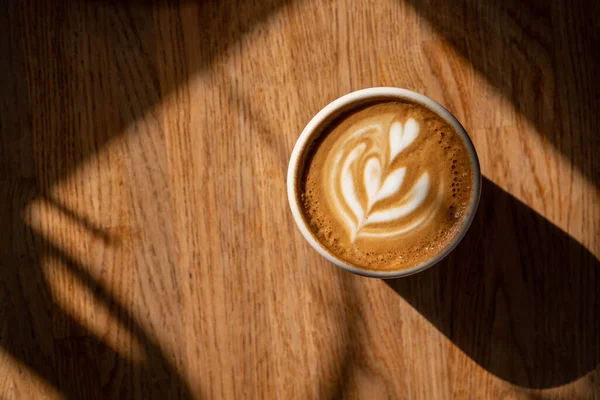 Secangkir kopi di atas meja kayu di bawah sinar matahari. Cappuccino segar dengan seni latte di latar belakang coklat. Busa yang indah. Konsep coffee break. Pagi minum dengan kafein. Tampilan atas, lay datar, ruang penyalinan — Stok Foto