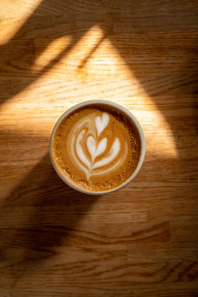 Cangkir kopi di atas meja kayu. Cappuccino segar dengan seni latte yang indah di latar belakang coklat dengan bayangan dan bintik-bintik matahari. Istirahat kopi. Foto atmosferik. Selamat pagi minum. — Stok Foto