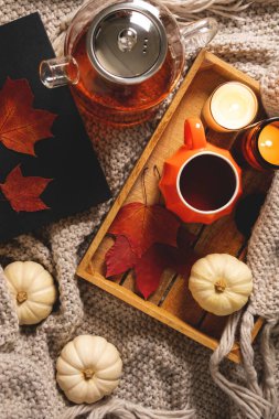Sonbahar kompozisyonu. Cam çaydanlık, bir fincan çay, kırmızı akçaağaç yaprakları, kitap, balkabağı ve bej ekoseli ahşap tepside mumlar. Karanlık atmosferik düşüş fotoğrafı. Yavaş bir yaşam tarzı. Üst manzara, düz la