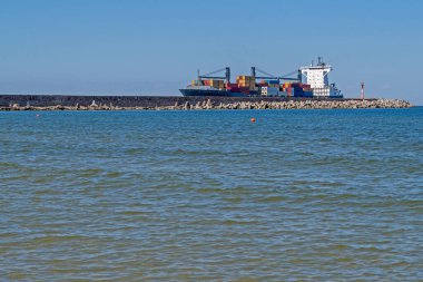 Farklı renklerde metal konteynırlarla dolu bir konteynır, kıyı koyuna girer. Mavi gökyüzüne karşı kırmızı ve beyaz bir deniz feneri ve deniz yüzeyine karşı gri bir taş iskeleyi geçer.