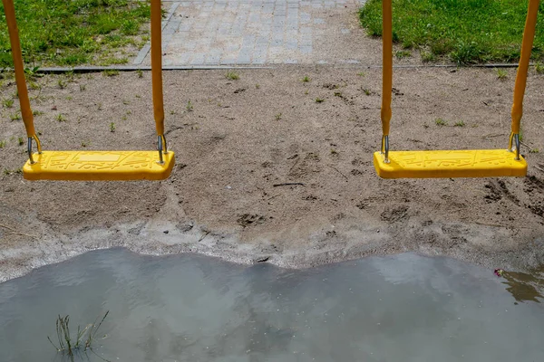 雨の後に形成された大きな水たまりの上にぶら下がっている2つの黄色のプラスチック製の懸濁液の振動 砂質土とその背後にある緑の芝生の間の舗装された歩道の背景に組成 — ストック写真