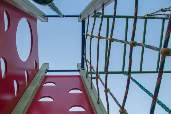 Construction on playground in form of tower, two walls of red wood with cutouts for climbing legs, third wall is grid of intertwined colored ropes, on fourth hangs thick green cable rope, bottom view