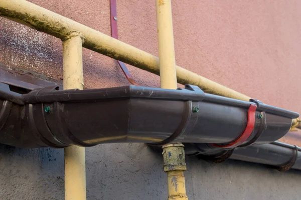 A fragment of a brown plastic after-rain drainage tray, curved with special fittings to encircle yellow gas pipes, mounted on the pink wall of the shabby facade of a residential building
