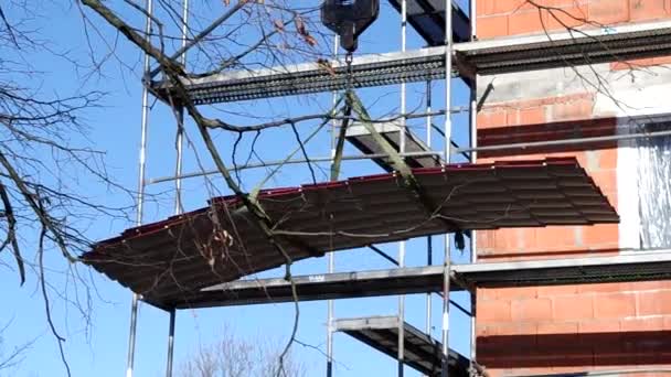 Lifting by black hook of red metal tiles roof section to upper level of brick house under construction, past mesh scaffoldings and growing nearby tree branches — Stock Footage Lifting Black Hook Red Metal Tiles Roof Section Upper Level Video Clip