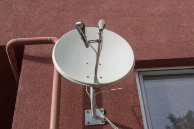 Pencerenin solundaki ve kemerin sağındaki bir apartmanın kahverengi cephesine çapa ile bağlı yuvarlak beyaz metal uydu TV anteni. Yakınlarda bir gaz boru hattı görünüyor..