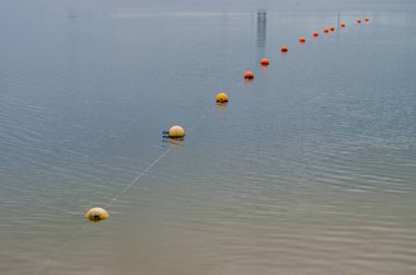 Sarı ve turuncu şamandıralar, kıyı şeridi boyunca küçük dalgalanmalarla kaplı yeşil masmavi su yüzeyine gerilerek gölün sığ sularını hapsettiler.