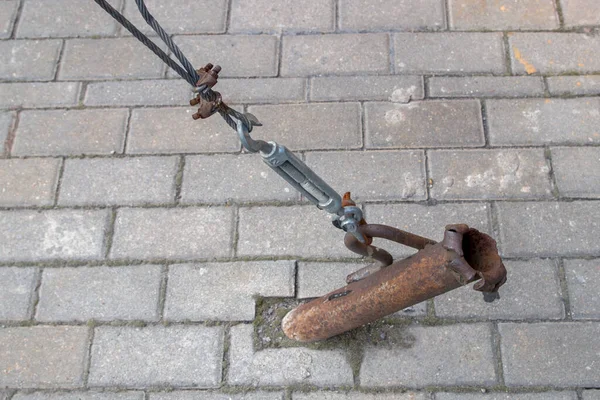 Stretching of metal rope by turnbuckle of ring-hook type attached to ear of rusty pipe embedded in ground and rusted cable clips on loop of steel multi-stranded cable on background of paving slabs